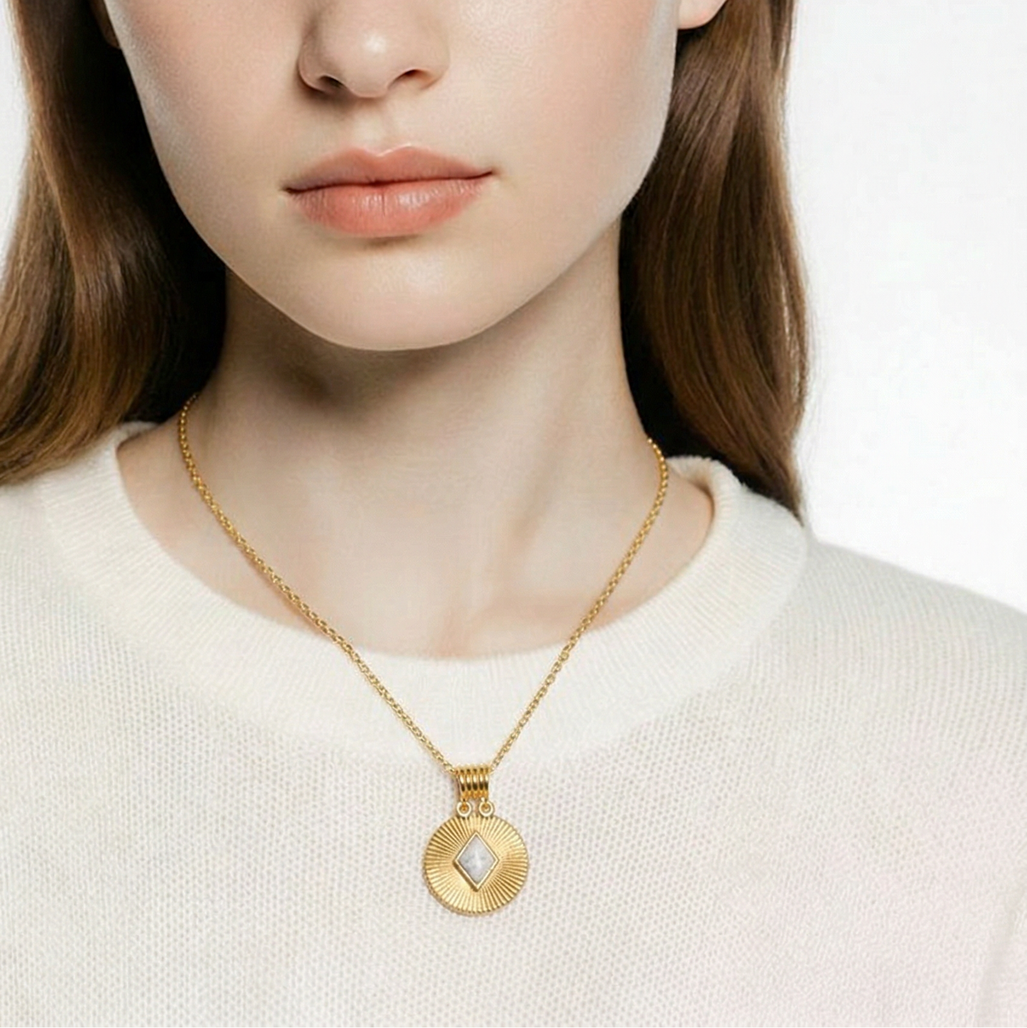 Woman wearing a gold necklace with a white  crew neckline t-shit on. Close-up of a person wearing a gold necklace with a circular pendant on a neutral background. The textured gold pendant has a white diamond shaped howlite gemstone in the middle of it.