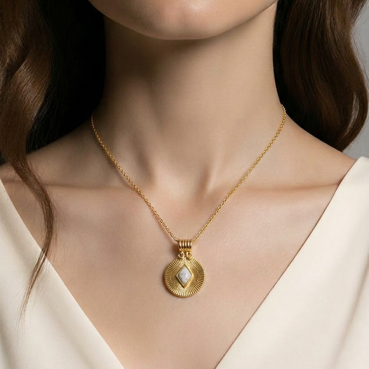 Close-up of a person wearing a gold necklace with a circular pendant on a neutral background. The textured gold pendant has a white diamond shaped howlite gemstone in the middle of it.