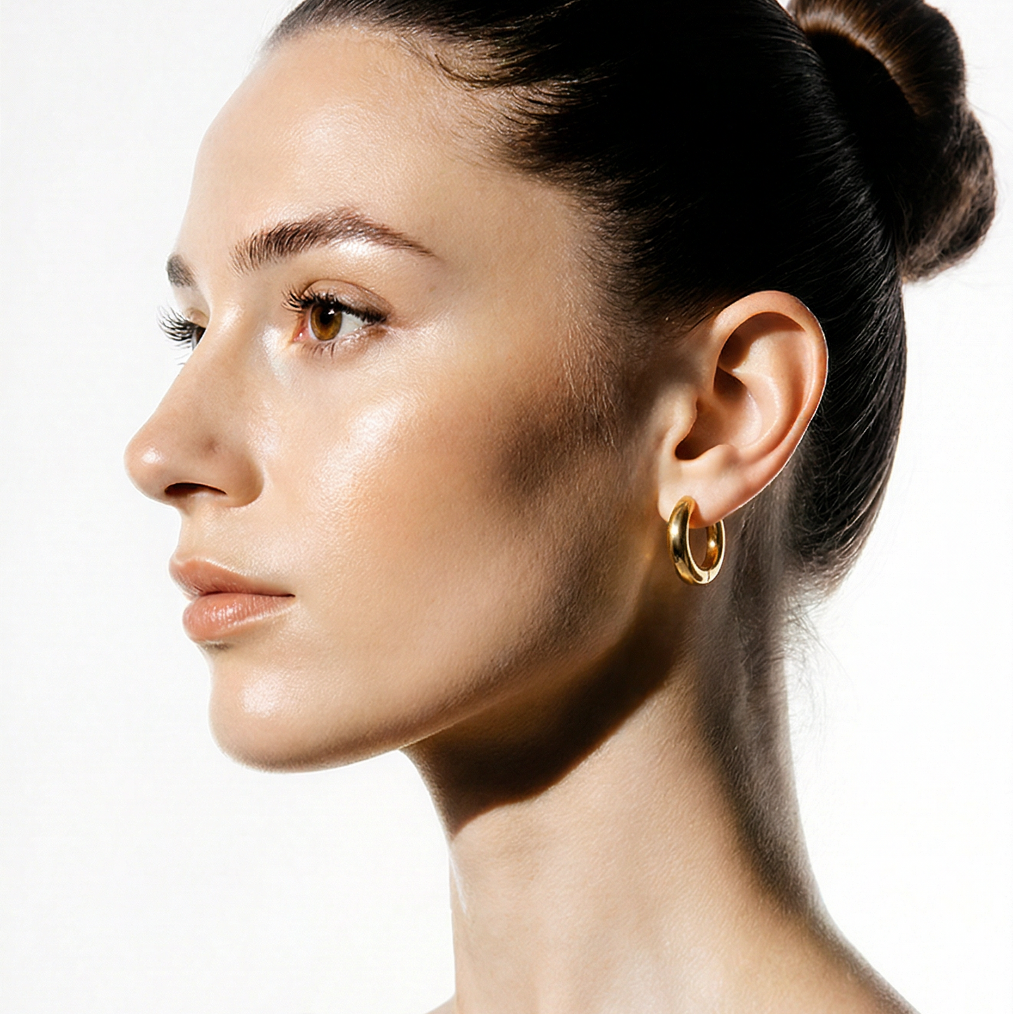 Woman with a bun hairstyle wearing gold chuncky hoop earrings on a white background