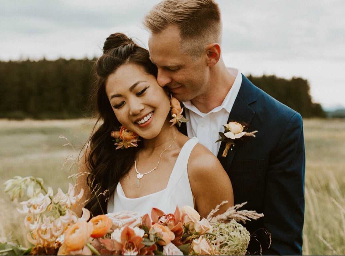 Bride and Groom embracing in a field with floral bouquets, bride wearing custom made gold necklace for her special day 